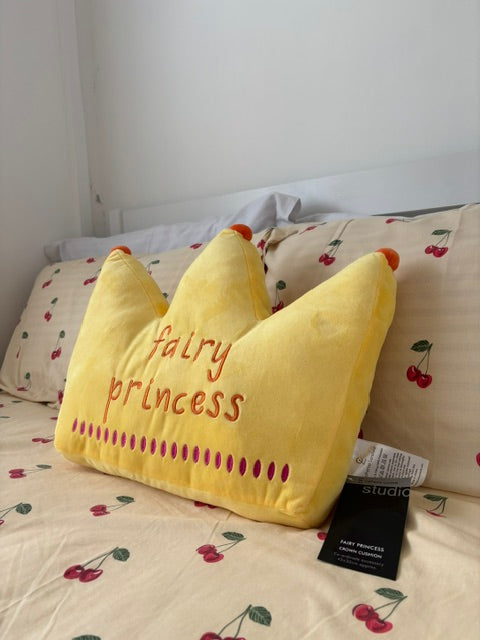 A yellow crown-shaped plush pillow with 'Fairy Princess' embroidered in red, resting on a cherry-patterned bedsheet next to its black product tag.