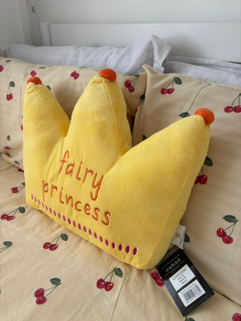 A yellow plush crown-shaped, embroidered with 'fairy princess' in red thread, resting on a cream-colored bedspread with a red cherry print. A black product tag is attached to the corner.