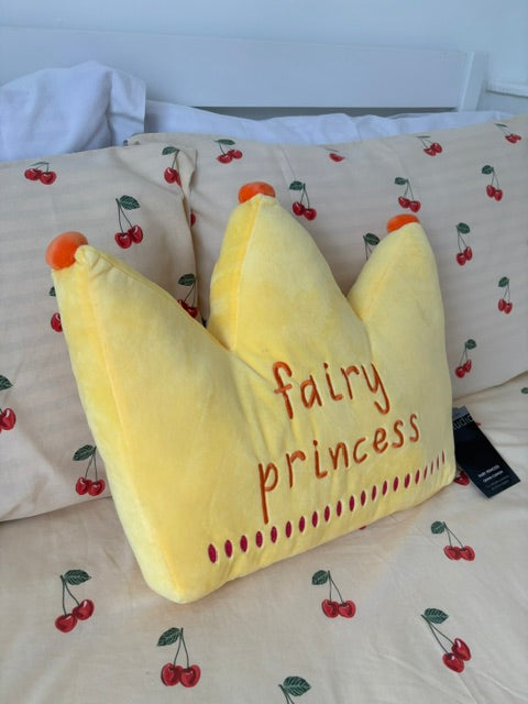 A yellow crown-shaped plush pillow with 'fairy princess' embroidered in red on the front, resting on a bed with cherry-patterned bedding.