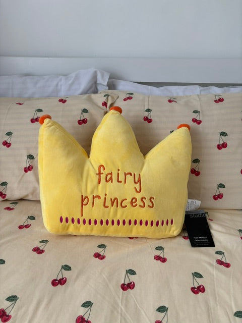 A yellow plush pillow shaped like a crown, embroidered with the words 'fairy princess' in red thread, resting on a bed with a cherry-patterned duvet.