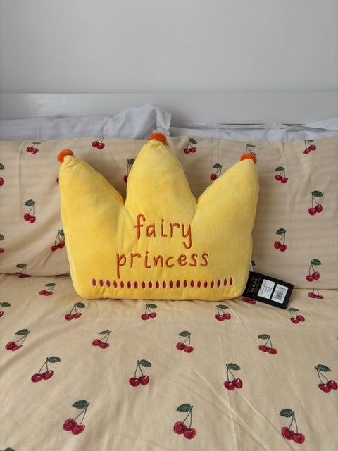 A yellow plush pillow shaped like a crown with the words 'fairy princess' embroidered in red, placed on a bed with a cherry-patterned beige duvet. A black product tag is attached near the top.