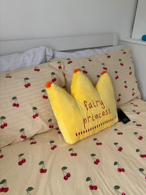 A yellow crown-shaped plush pillow with 'fairly princess' embroidered on it, resting on a bed with cherry-patterned bedding.
