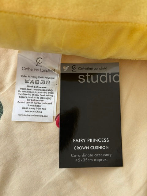 A close-up of a black product tag reading 'FAIRY PRINCESS crown cushion' and a white sewn-in care label for Catherine Lansfield, placed on a cream-colored fabric next to a yellow fabric.
