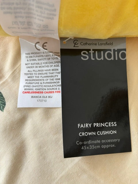 A black fabric tag labeled 'FAIRY PRINCESS CROWN CUSHION' and 'studio' by Catherine Lansfield, placed on a cream-colored textured fabric surface next to a yellow item.