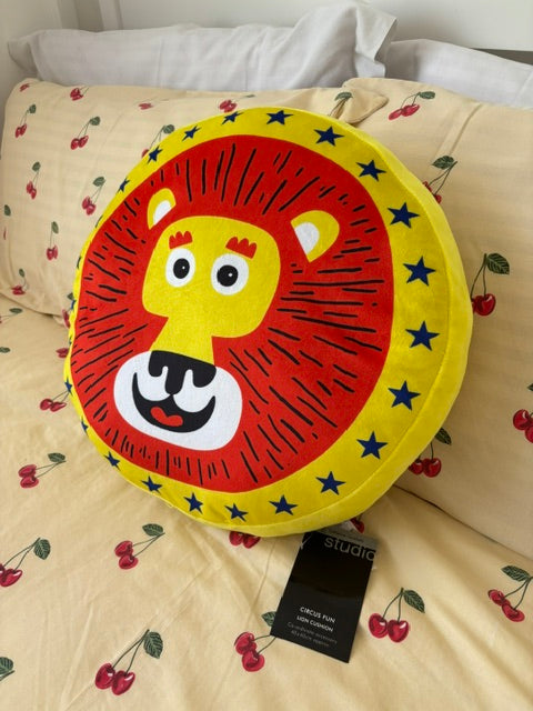 A round lion-face throw pillow with a red and yellow design, placed on a beige cherry-print bedsheet next to a black tag.