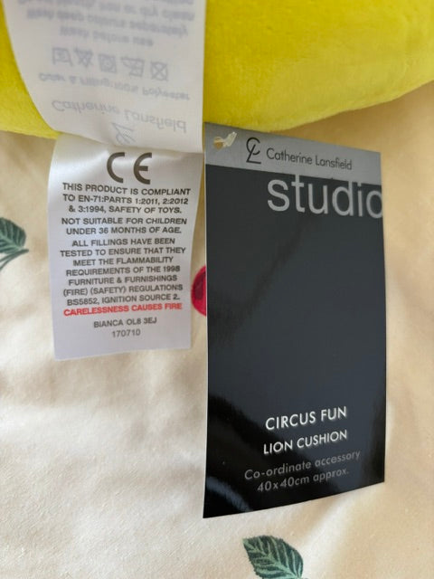 A close-up of a black fabric tag reading 'studio' and 'CIRCUS FUN LION CUSHION' with product details, alongside a white care/information label and part of a yellow plush item on a light fabric background with green leaf print.