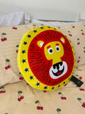 A round lion-face cushion or pillow with a red mane, yellow face, and blue stars, placed on a cream-colored bedspread with a cherry print.