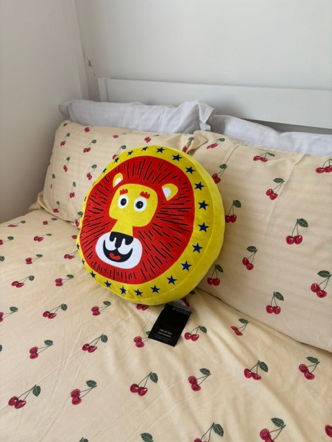 A round plush lion-face pillow with a red lion mane, yellow face, and black star border, resting on a cream-colored bedspread printed with red cherries.