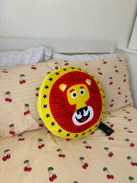 A round lion-face throw pillow in red, yellow, and white, placed on a bed with cream-colored bedding printed with red cherries.