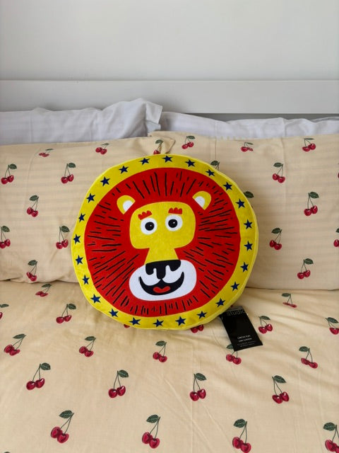 A round lion-face decorative pillow with a red mane and yellow face, placed on a cream-colored bedspread printed with red cherries.