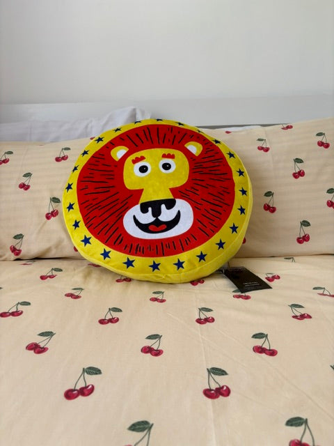 A round lion face throw pillow with a red and yellow mane, placed on a cream-colored bedspread with a red cherry print.