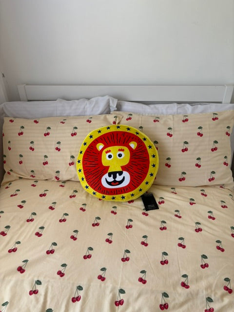 A round lion-face throw pillow with a red and yellow mane, placed on a cream-colored bedspread printed with a red cherry pattern.