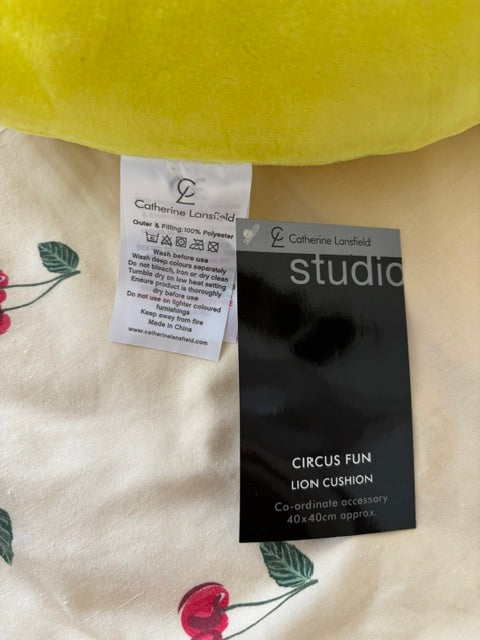 A black product tag for the 'studio Circus Fun Lion Cushion' by Catherine Lansfield lies on a cream-colored fabric with a cherry print pattern, next to a yellow plush item.