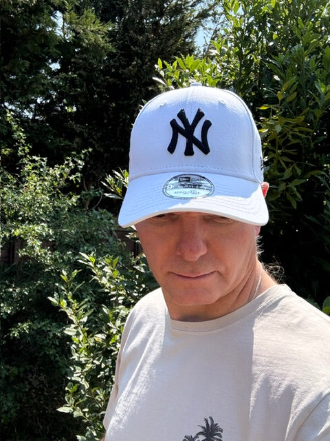 A man wearing a white New York Yankees baseball cap and a white t-shirt, photographed outdoors against a background of green trees.