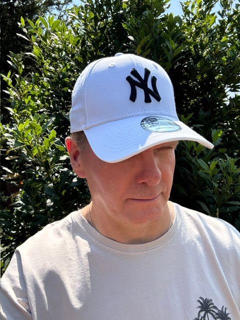 A man wearing a white New York Yankees baseball cap and a white t-shirt, photographed outdoors against a background of green foliage.