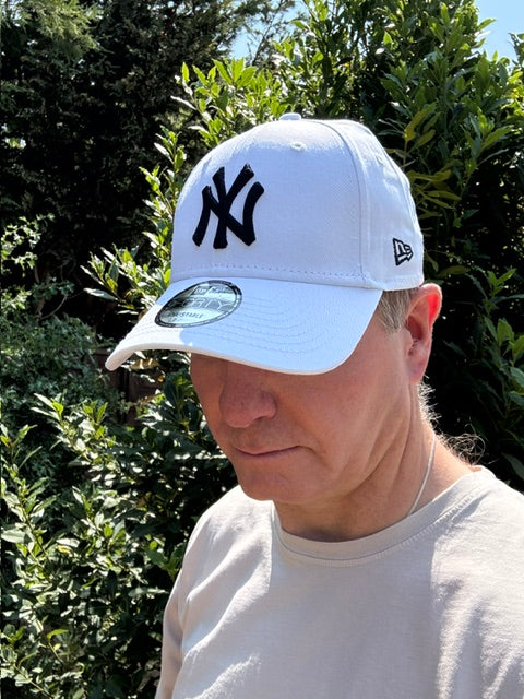 A white New Era baseball cap with a black New York Yankees logo, worn by a person outdoors in front of green foliage.