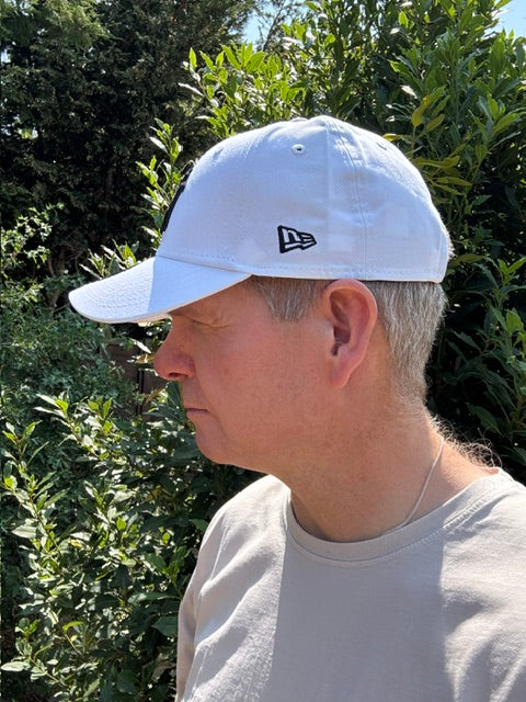 A man wearing a white New Era baseball cap and a light gray t-shirt, photographed outdoors against a background of green foliage.