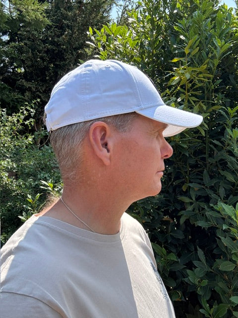 A man wearing a white baseball cap and a white t-shirt, photographed outdoors against a background of green foliage.