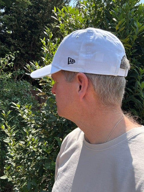A man wearing a white New Era baseball cap and a light gray t-shirt, photographed outdoors against a background of green foliage.