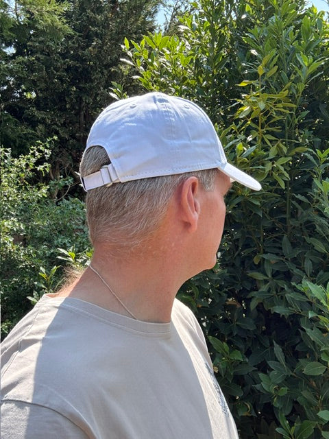 A man wearing a white baseball cap and a light gray t-shirt, photographed outdoors against a background of green trees.